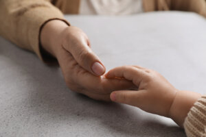 Woman holding hands with her little daughter at light grey table, closeup